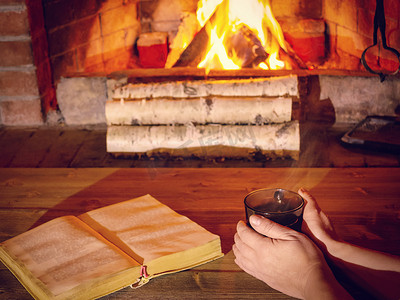 女人的手在燃烧的壁炉旁的一杯热茶上暖和起来，桌上放着一本打开的书
