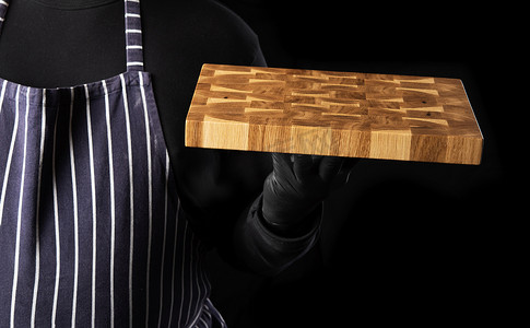 穿着蓝色条纹围裙和黑色衣服的男厨师站着，手里拿着一块长方形的木制厨房砧板