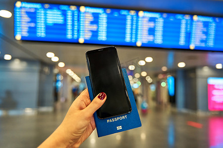 手机店招摄影照片_在机场的信息板背景下，一只手拿着智能手机和出国旅行的护照