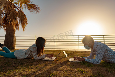 两名学生在日落时躺在草地上使用笔记本电脑在热带海洋度假胜地的城市公园学习课程。