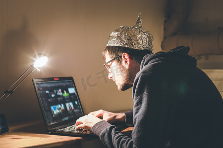 阴谋论概念：戴着铝盖的年轻人上网搜索，孤独地坐在黑暗的地下室里