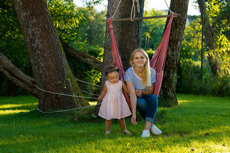 母亲带小孩摄影照片_一位带着小孩的年轻妇女坐在她花园里的吊床上。
