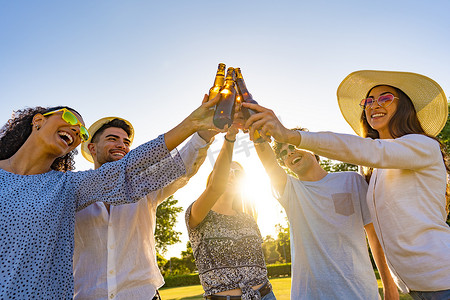 一群年轻的时髦朋友在日落前庆祝户外喝啤酒和用瓶子敬酒。