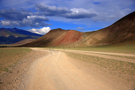 沙漠地区摄影照片_漫天雷云下，土路蜿蜒在红色山脚下。