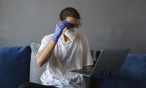 隔离期间，戴着医用口罩和橡胶手套的男子坐在家里，在沙发上用笔记本电脑工作。
