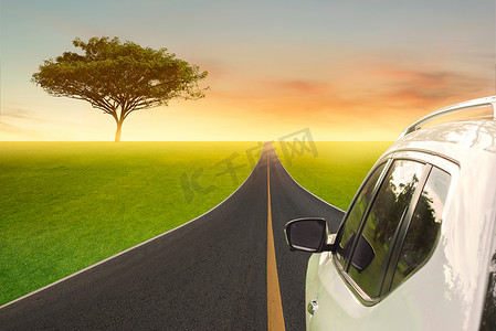 一辆白色的汽车在乡村的柏油路上行驶，与云彩和美丽的夕阳映衬着绿色的田野。