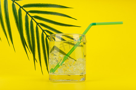 用棕榈叶装饰的黄色背景上装满冰块的苏打水、柠檬水或冷清凉饮料的玻璃