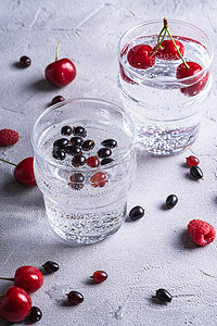 石材混凝土背景上两块透明玻璃杯中含樱桃、覆盆子和醋栗浆果的新鲜冷苏打水饮料、夏季饮食饮料、角度宏观