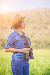 手持设备摄影照片_女人戴帽子，在草地上拿着双筒望远镜