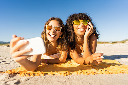 两个戴着太阳镜的可爱女孩在暑假使用智能手机躺在热带海滩的海沙上自拍的正面肖像。
