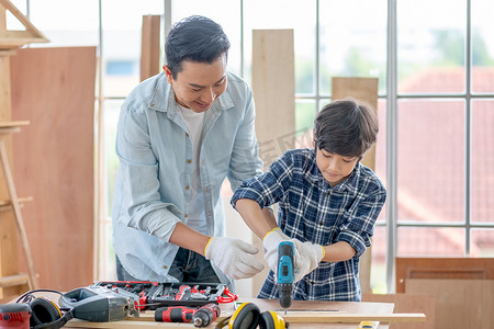 电钻摄影照片_亚裔父亲教他的儿子使用电钻在家里做木工。