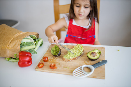 营养膳食餐摄影照片_餐桌旁的小女孩在一片面包上撒鳄梨