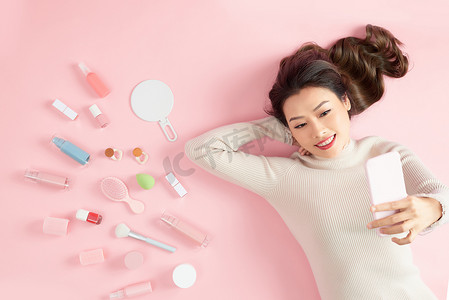 年轻的亚洲女人用手机自拍，躺在粉红色的背景上，拿着化妆品。