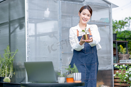 植物产品摄影照片_美丽的亚洲女孩拿着植物产品，对着镜头展示，也站在温室工作区前