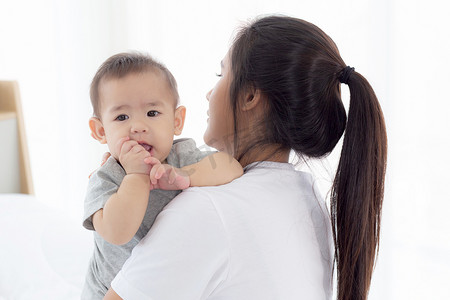 年轻的亚洲母亲在家里的卧室里抱着小女婴，妈妈带着孩子和照顾孩子，女人和孩子无忧无虑，蹒跚学步的孩子和父母，情感和表达，家庭观念。