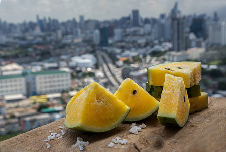 新鲜多汁的黄色西瓜在阳台的旧木桌上切成三角形，可欣赏城市景观。