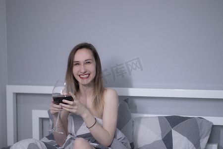 快乐美丽苗条的半裸女孩在床上拿着一大杯红酒，床上放着三角形的灰色床单