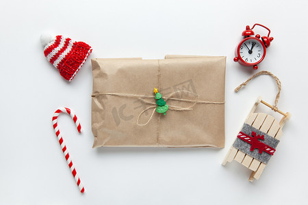 用棕色牛皮纸包裹的圣诞礼物，用鞭子绑着，上面有甘蔗糖、小模拟钟、雪橇、白色背景的帽子。