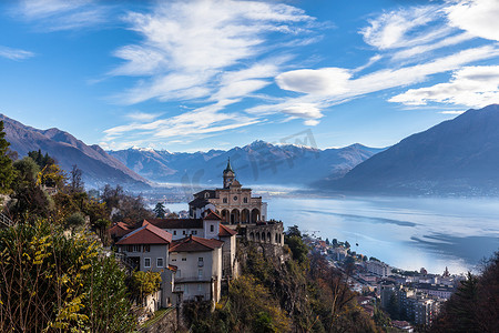 瑞士提契诺州秋季背景中洛迦诺市上方的麦当娜德尔萨索教堂与马焦雷湖、白雪覆盖的瑞士阿尔卑斯山峰和蓝天云的壮丽空中全景