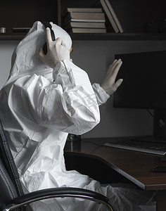 身穿医用口罩和防护服、戴手套的男子坐在家里，在隔离期间与桌上的电脑一起工作，并向他的商业伙伴或朋友问好。