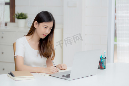 年轻的亚洲女性在笔记本上写作以计划在家里的办公桌上工作和使用笔记本电脑，关于金融、女性学习和学习、商业和沟通概念的女孩笔记。