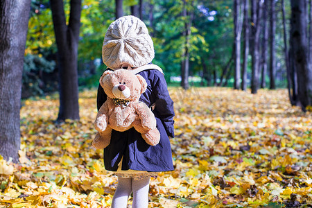 婴儿背包摄影照片_秋日阳光明媚的森林里背着背包熊的迷人小女孩的后视