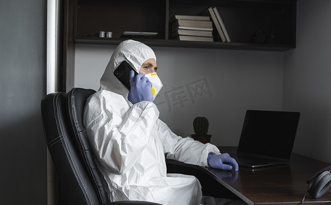 身着防护服、医用口罩和橡胶手套的男子坐在家里，在隔离期间与朋友或商业伙伴通电话。