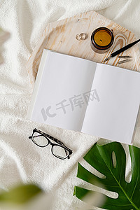书书样机摄影照片_打开的书、眼镜、蜡烛和鲜花在白色床上的顶视图。