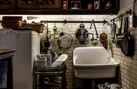 厨房里有工具、平底锅、挂在砖白墙上的锅和放在电线架上的玻璃瓶、放在冰箱和陶瓷浴缸上的木托盘。