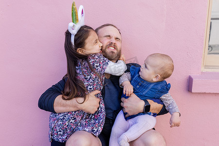 粉色墙壁摄影照片_快乐的父亲抱着两个可爱的女儿，背景是粉红色的墙壁