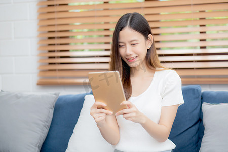 年轻迷人的亚洲女性在家里的沙发上使用浏览平板电脑休息，快乐的女孩坐在沙发上放松阅读家里的数字小工具、交流和生活方式概念。