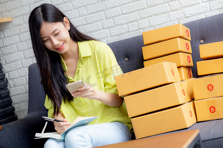 年轻的亚洲女性是企业家，他们在家里用笔记本电脑在线查看电话和写客户订单、网上购物、物流和送货，女性是在沙发上工作的卖家，商务中小企业。