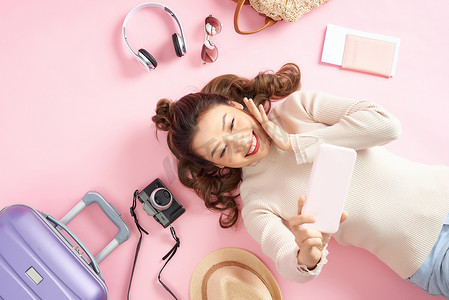 旅游行李卡片摄影照片_快乐的亚洲女孩用手机拍照，带着旅行行李躺在粉红色的地板上。