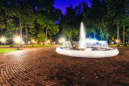夜间公园里有一个喷泉，水流模糊，灯笼照亮，铺着石头路面、树木和长椅。