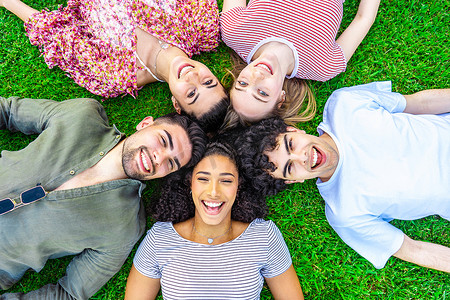 五个快乐的年轻多种族 z 世代朋友头对头躺在公园草坪的草地上，从底部看着镜头。