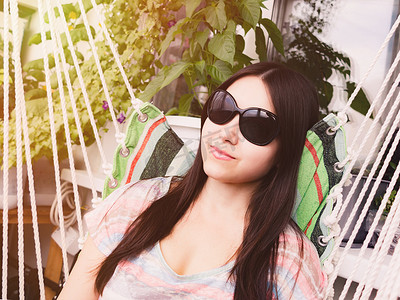 戴着墨镜、长着深色头发的漂亮女孩坐在阳台的吊床上休息