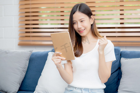 年轻迷人的亚洲女性在家里的沙发上使用浏览平板电脑休息，快乐的女孩坐在沙发上放松阅读数字小工具，在家里兴奋、交流和生活方式概念。