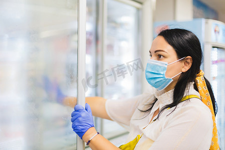 戴着防护面罩和一次性手套的女性在超市购物时浏览冷冻食品区，有选择地关注