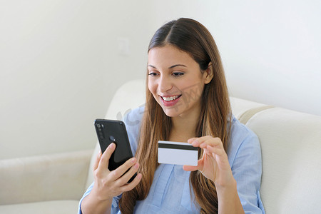 女士坐在家里客厅的沙发上，用信用卡和智能手机在网上购物