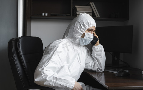 戴着医用口罩和橡胶手套的男子坐在家里，在隔离期间与朋友或商业伙伴通电话。