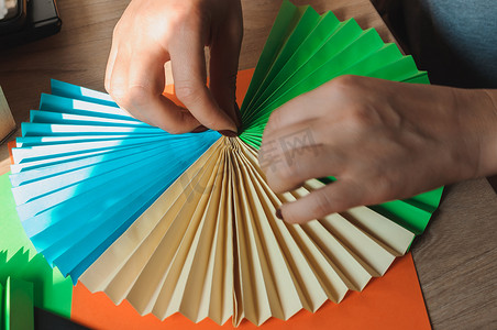 在前景中，女性手在木桌上粘着折纸扇、彩色纸片和剪刀。