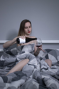 美丽苗条的半裸女孩把红酒从没有标签的瓶子里倒进一个巨大的玻璃杯里，躺在床上，床上铺着三角形的灰色床单