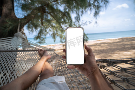 模拟手机摄影照片_手拿着白色场景的模拟手机，躺在海滩上树木之间的吊床上秋千上。