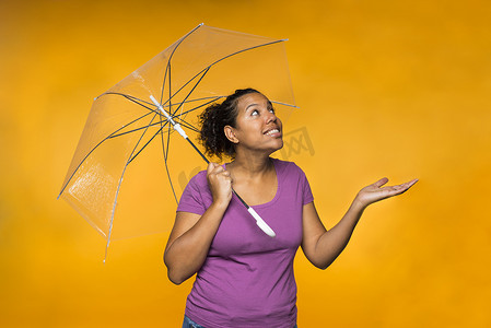 下雨水摄影照片_黄色背景下穿着紫色衬衫的年轻漂亮混血女性拿着雨伞检查雨水