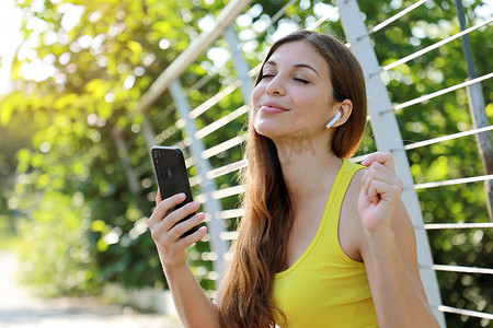 一个快乐的年轻健身女孩坐在公园里享受使用带无线耳机的手机听音乐的清晨肖像