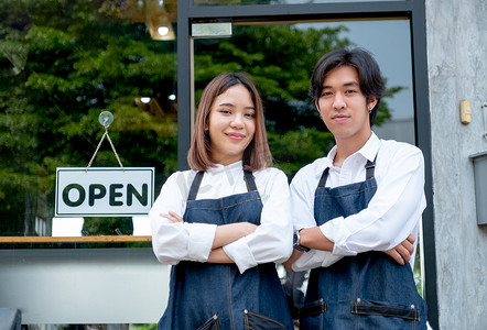 两个亚洲咖啡师或咖啡师男人和女人双臂交叉或自信地站着，也看着镜头，在咖啡店前微笑。