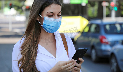 COVID-19 大流行性冠状病毒移动应用程序-戴着外科口罩的年轻女性在城市街道使用智能手机应用程序帮助追踪接触者以应对 2019-20 冠状病毒大流行