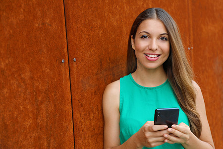 微笑的漂亮年轻女人穿着便装站在锈迹斑斑的建筑物墙上，用智能手机发短信，看着相机。
