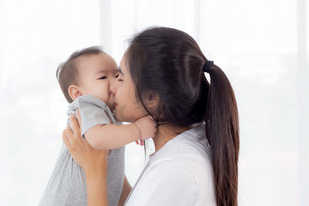 年轻的亚洲母亲抱着小女婴在家里的卧室里一起亲吻，妈妈带着孩子和照顾，女人和孩子无忧无虑，蹒跚学步的孩子和父母，情感和表达，家庭观念。