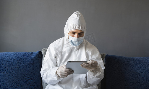 身着防护服、医用口罩和橡胶手套的男子坐在家里，在隔离期间在沙发上使用平板电脑工作。
